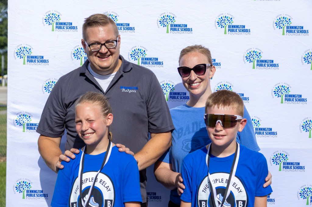A smiling family posing together in front of a backdrop featuring the Bennington Public Schools Foundation logo. The group includes two children wearing medals, a man in a blue branded shirt, and a woman with sunglasses.