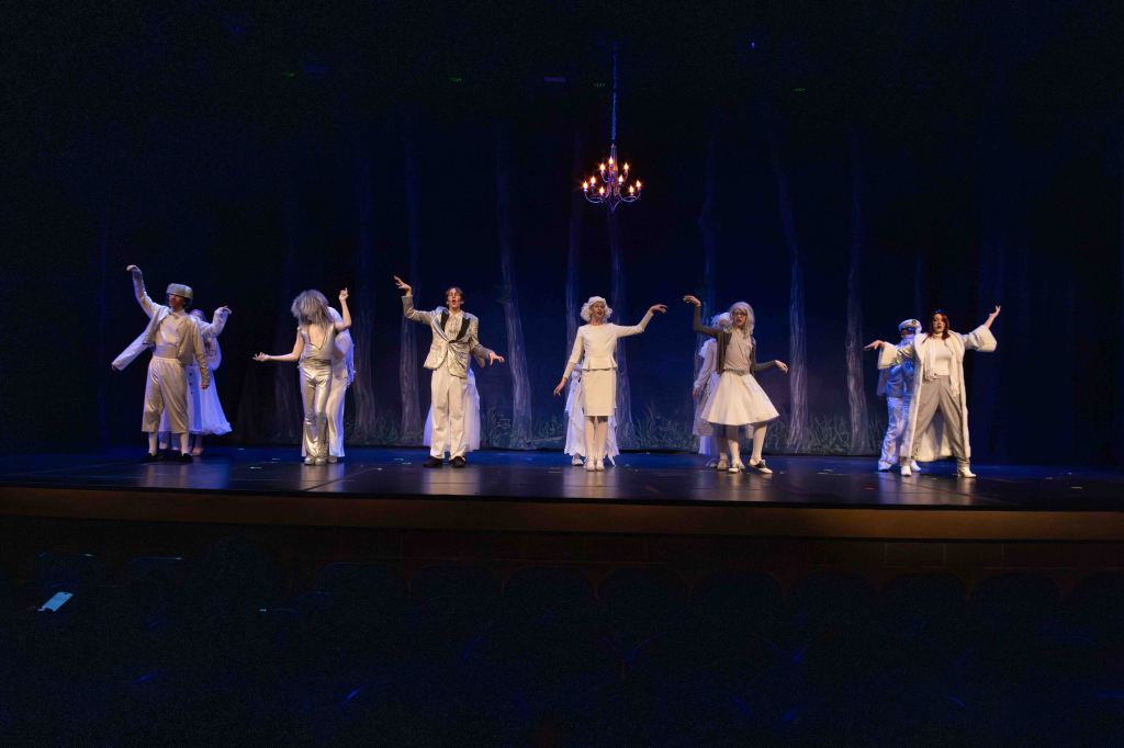 A theatrical performance featuring eight actors dressed in white costumes, dancing on stage under a chandelier, with a dark forest backdrop.