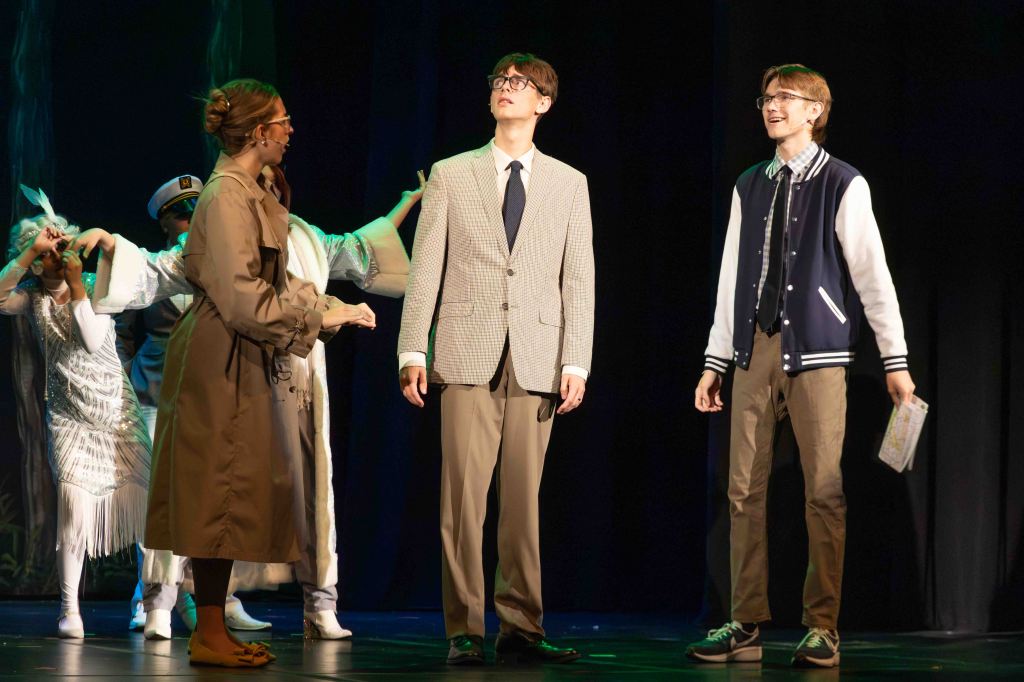 A theatrical performance scene featuring three characters on stage. Two young men are dressed in formal attire, one in a suit and the other in a casual varsity jacket, while a woman in a trench coat engages with them. In the background, additional performers in costumes are partially visible.