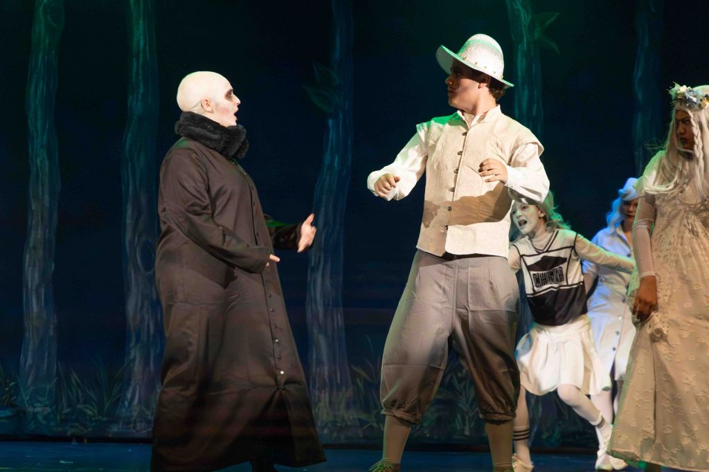 Two performers in a theatrical production, one dressed in a long black coat and bald cap, the other in a light-colored vest and hat, engaged in a dramatic conversation on stage with other characters in the background.