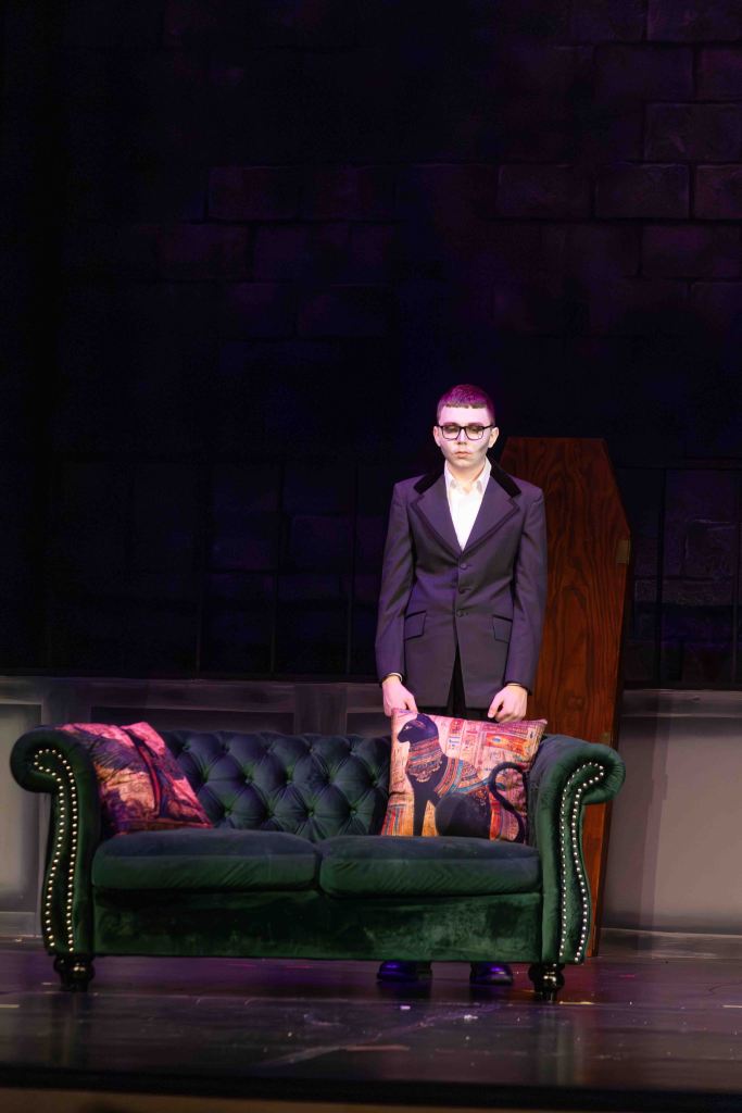 A person in a tuxedo stands on stage beside a green velvet couch adorned with decorative pillows, against a dark brick background.
