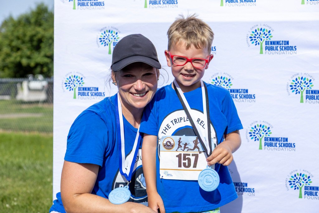 A smiling woman and a boy with glasses, both wearing medals, pose together in front of a backdrop featuring the Bennington Public Schools Foundation logo.