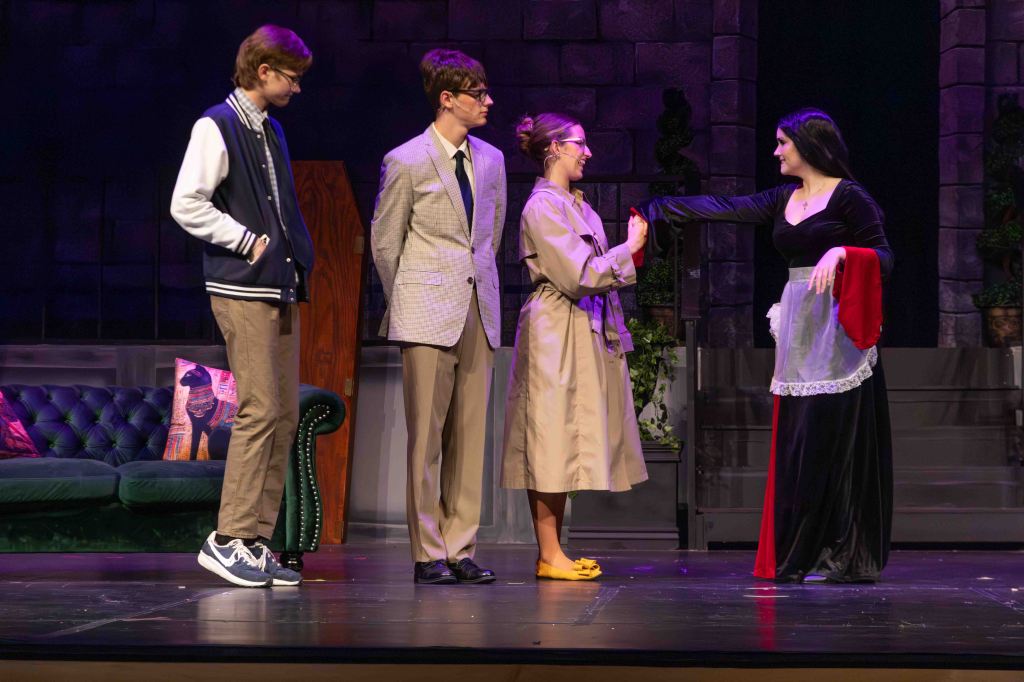 A scene from a stage play featuring four characters: two in casual attire, one in a trench coat, and one in a black dress with a red accent, interacting on a theatrical set with a couch and decorative plants in the background.