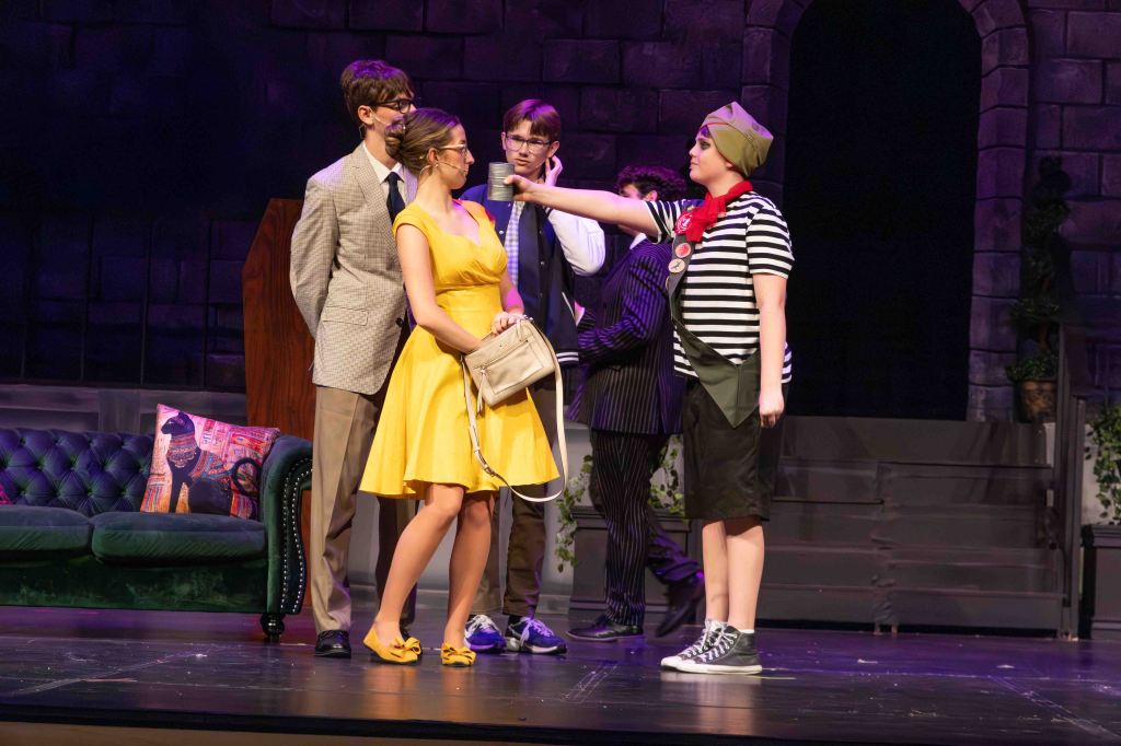 A scene from a theatrical performance featuring five actors in various costumes. A woman in a yellow dress stands center stage, holding a bag, while a girl in a striped shirt and green sash gestures towards her. The backdrop includes a decorative couch and a stone wall.