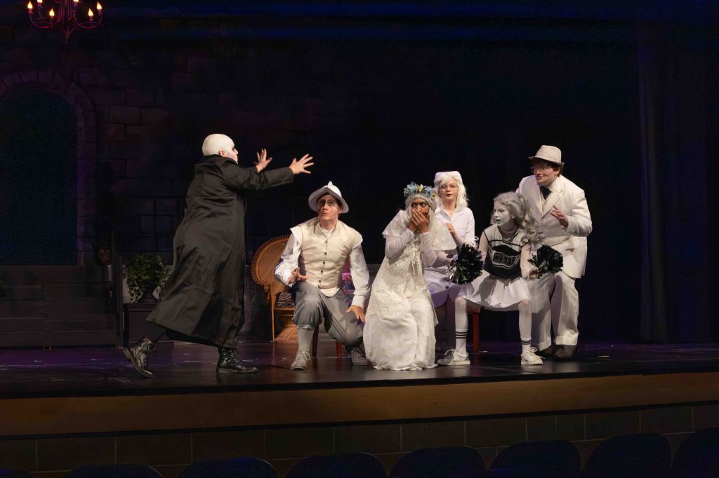 A theatrical performance featuring actors in various costumes, including a figure in a dark cloak, a bride in white, and characters in cheerleader outfits. They are gathered on stage with a dramatic backdrop.