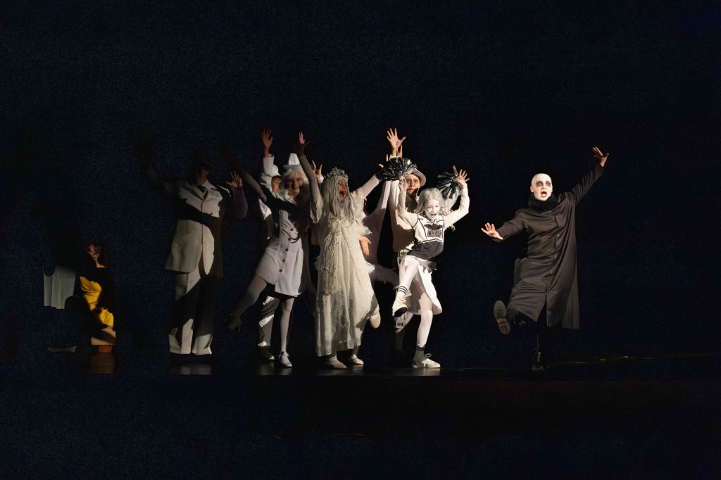 A theatrical performance featuring six performers in dramatic costumes, expressing excitement and energy on stage in front of a dark background.
