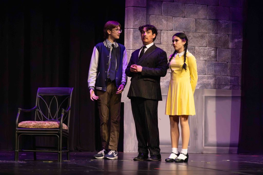 Three actors on stage during a theatrical performance, featuring a young man in a varsity jacket, a man in a pinstripe suit, and a young woman in a yellow dress with braided hair.