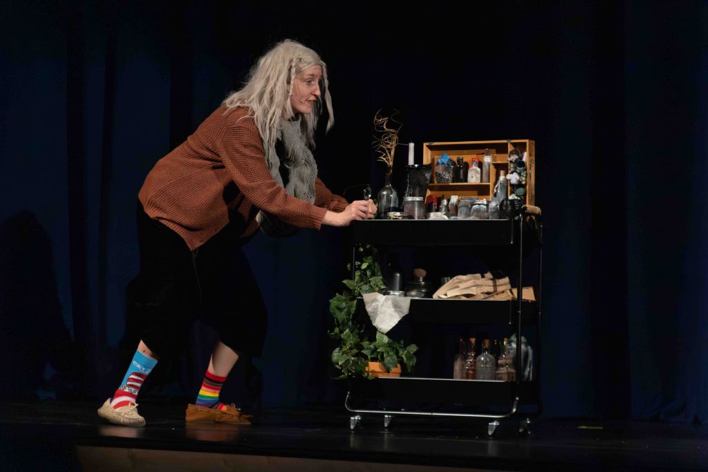 A performer dressed as an elderly character with long gray hair, wearing a cozy sweater and colorful socks, is interacting with a cart filled with various bottles and props on stage.