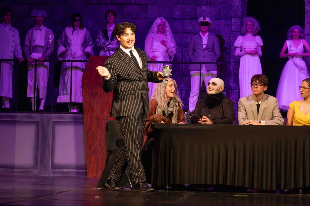 A performer in a pinstripe suit gestures while holding a mug on stage, surrounded by a mix of characters including a ghostly figure, a woman with long hair, and others seated at a table during a theatrical performance.