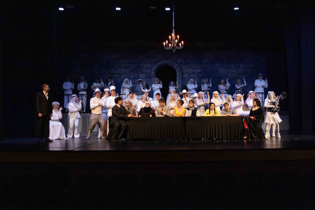 A theatrical performance featuring a large cast on stage, with actors in varied costumes including white attire and formal wear, gathered around a long table with a dark tablecloth under a chandelier.