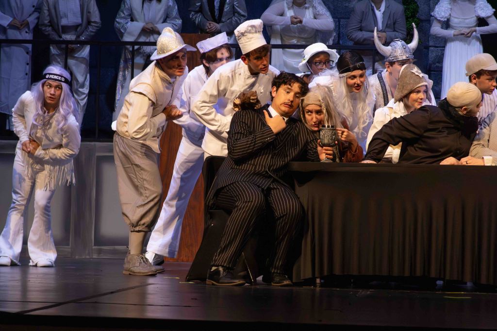 A scene from a theatrical performance featuring several actors in costumes, including a man in a black striped suit seated at a table, surrounded by other characters in varying outfits, engaged in a comedic display.