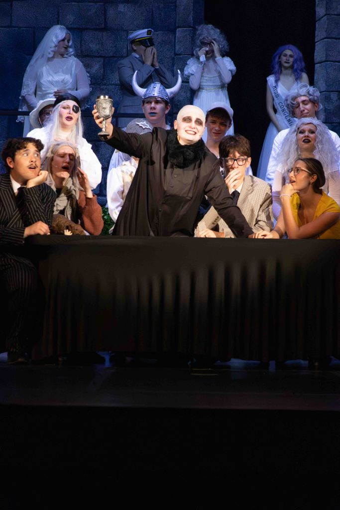 A theatrical scene featuring a cast of characters in dramatic costumes, with one central character raising a goblet in a celebratory pose. The background includes various figures in costumes such as brides and a military officer, while others are engaged in different emotional expressions.