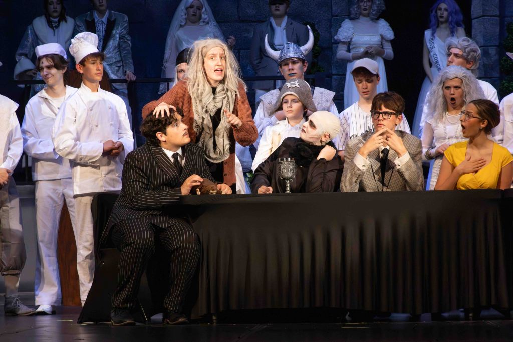 A theatrical scene featuring a diverse cast of characters on stage. In the foreground, actors are seated at a table, displaying various expressions. One actor with gray hair passionately gestures, while others observe intently. The background shows additional characters dressed in costumes, contributing to the dramatic atmosphere of the performance.
