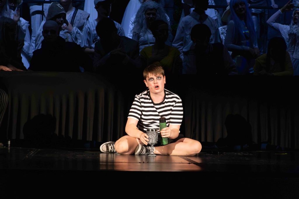 A young performer sitting on stage, wearing a striped shirt and holding a green object, with theatrical makeup. In the background, an audience is partially visible.