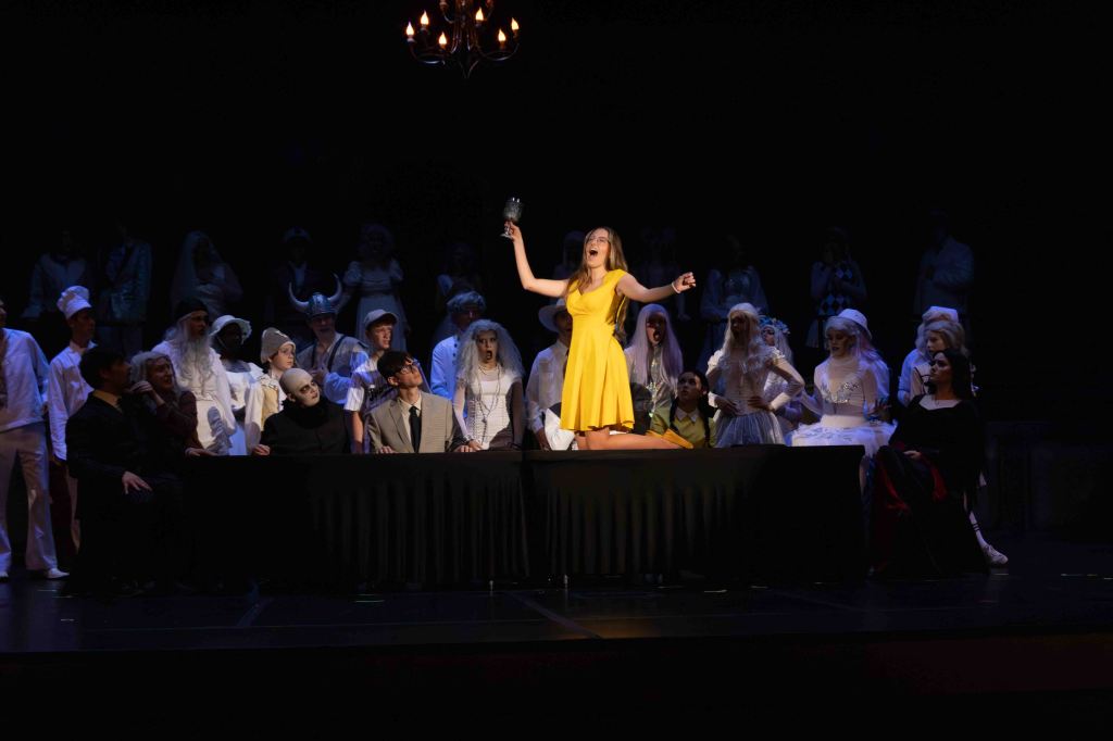 A performer in a bright yellow dress passionately sings on a stage, standing on a table surrounded by a diverse group of characters in costumes, while the audience watches.