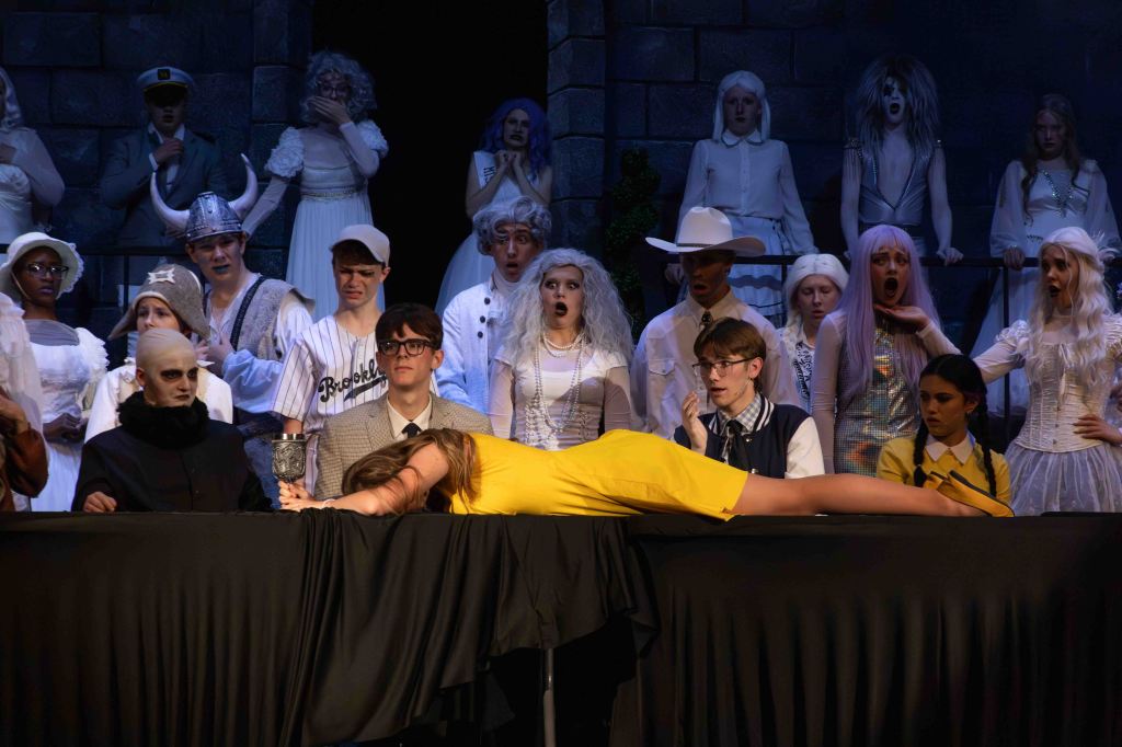 A dramatic stage scene featuring a group of actors in various costumes. In the foreground, a woman in a yellow dress lies on a table, while several other characters look on with expressions of surprise and concern. The background includes actors in costumes resembling a ghostly ensemble.