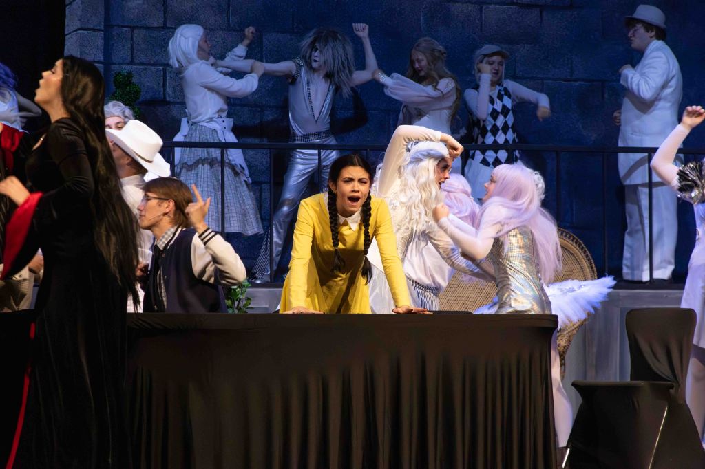 A lively theater scene featuring multiple characters in dramatic costumes, with one woman in a yellow dress expressing emotion at the forefront. The background includes other actors engaged in various actions, highlighting a chaotic or expressive moment.