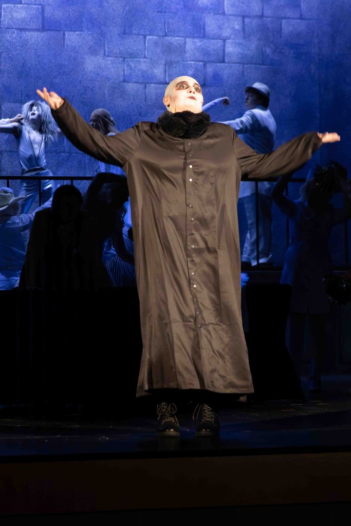 A performer in a dark, long coat and dramatic makeup stands confidently on stage with arms raised, surrounded by a theatrical set.