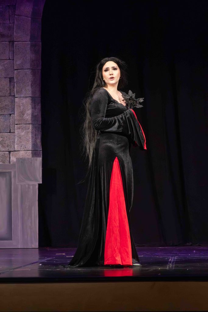 A performer in a black and red dress stands on stage, looking dramatic with long dark hair and holding a black prop.