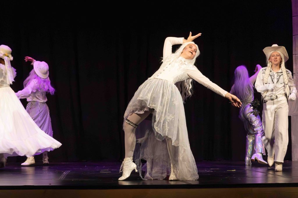 A group of performers on stage showcasing a dance routine. One performer in a white dress strikes a dramatic pose while others in vibrant outfits appear in the background.
