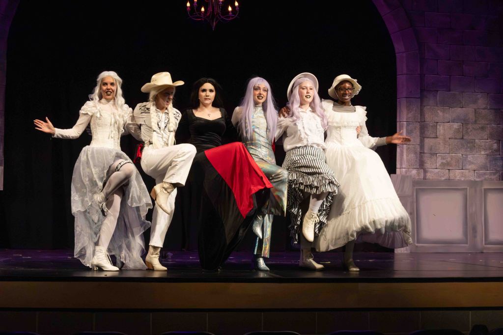 A group of six performers in theatrical costumes poses on stage, showcasing a variety of elaborate outfits, including white dresses, a black gown, and a colorful suit. They are smiling and striking playful poses, with a stone wall backdrop and stage lighting.