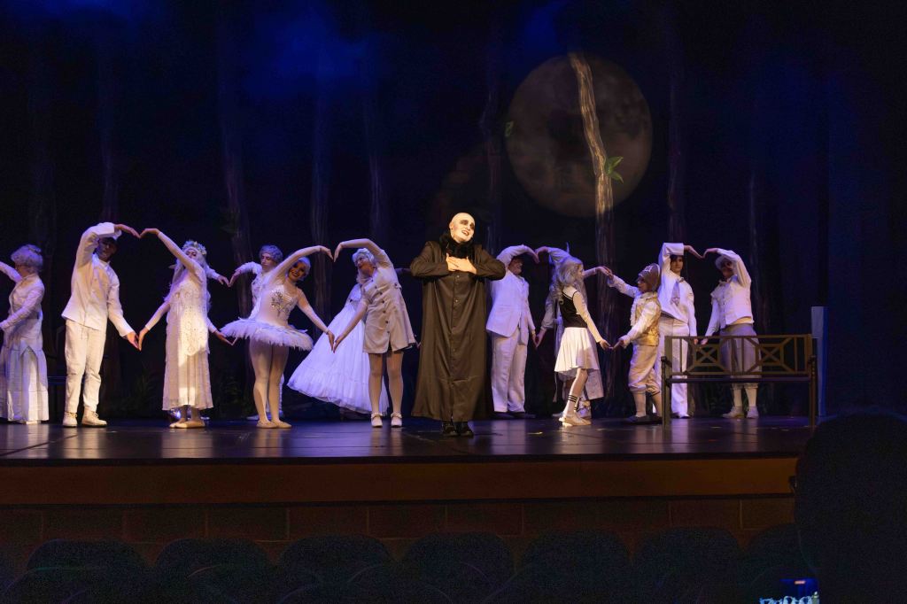 A theatrical performance featuring a group of dancers in white costumes and one performer in a black cloak, all on a stage decorated with a large moon backdrop.