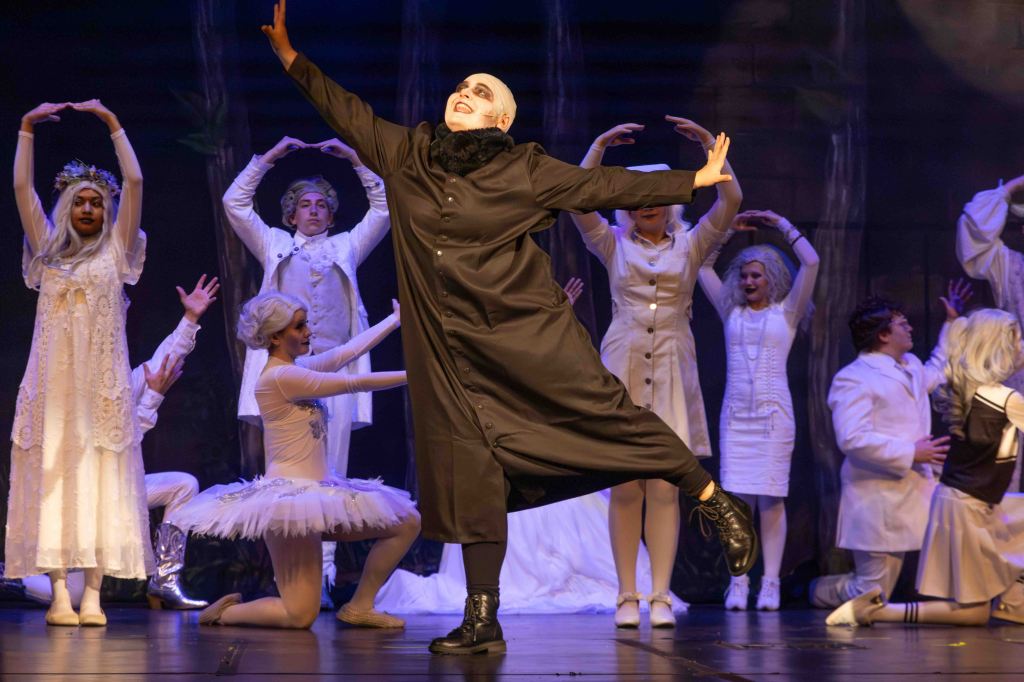 A theatrical performance featuring a dynamic character in a black costume posing dramatically, surrounded by dancers in white costumes, including a ballerina and figures in elaborate outfits, all on a stage with a forest backdrop.