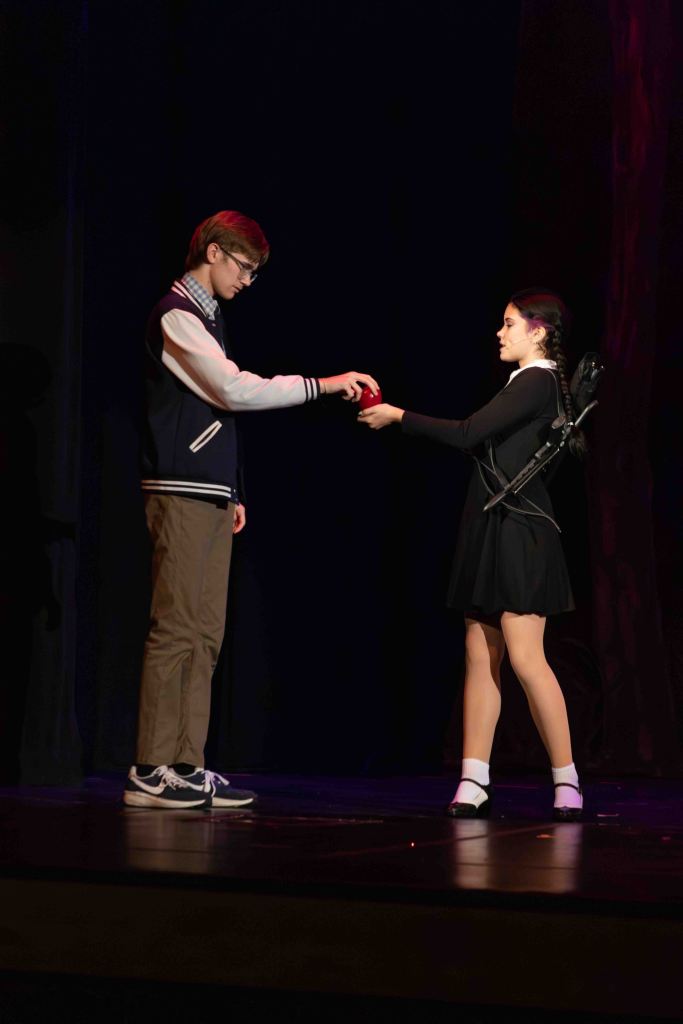 A boy in a varsity jacket hands a red object to a girl in a black dress on stage, illuminated by soft lighting.