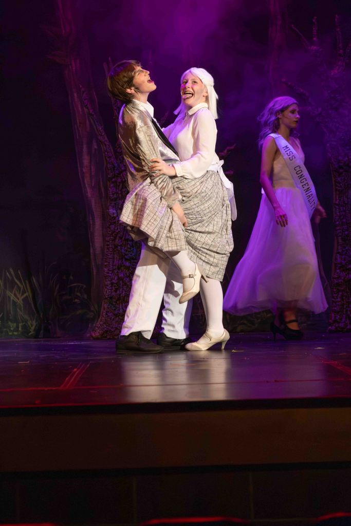 A theatrical performance featuring two actors in costumes expressing joy on stage, with a background of stylized trees and smoke effects.