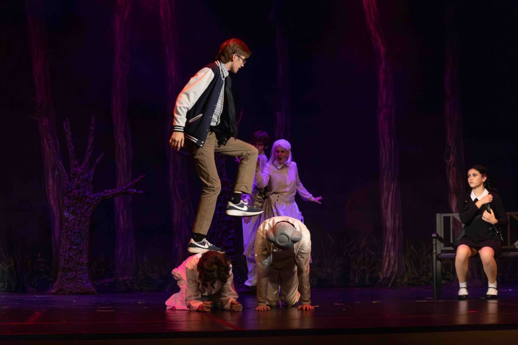 A theatrical performance scene featuring a boy jumping on another actor's back, surrounded by three other performers in a dark, forest-like setting.
