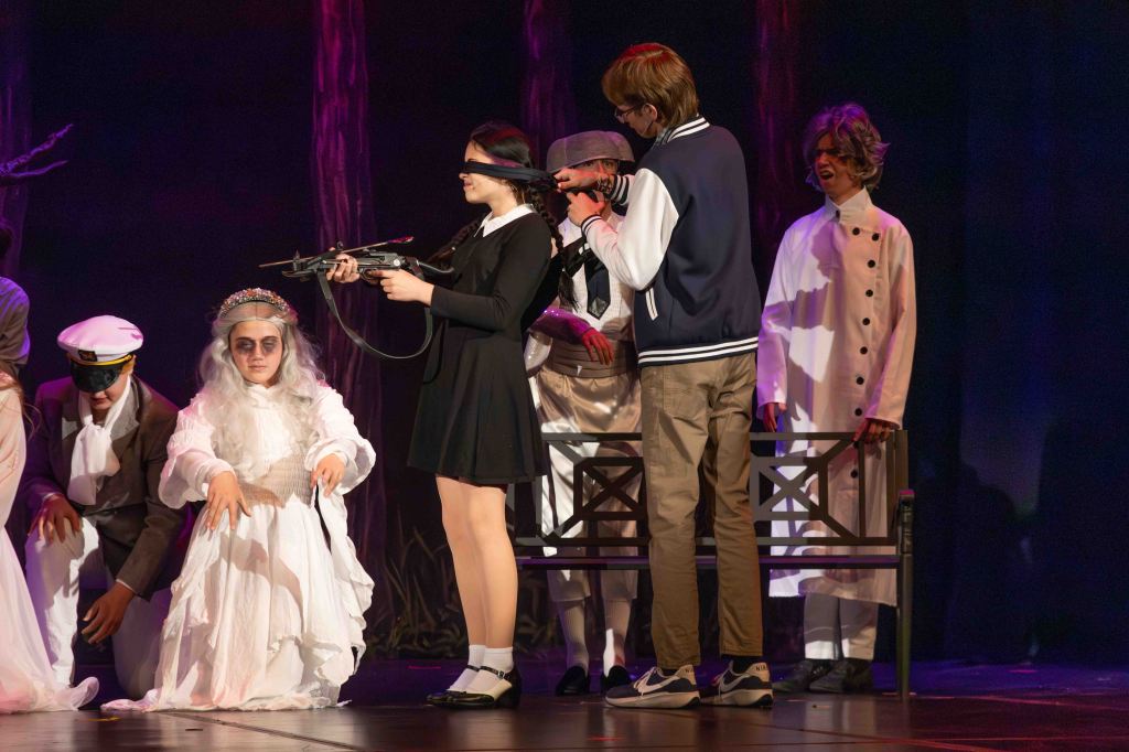 A theatrical scene featuring several actors performing on stage. A blindfolded female character in a black dress holds a prop firearm, while other characters in various costumes observe. The background includes dark trees, creating a dramatic atmosphere.