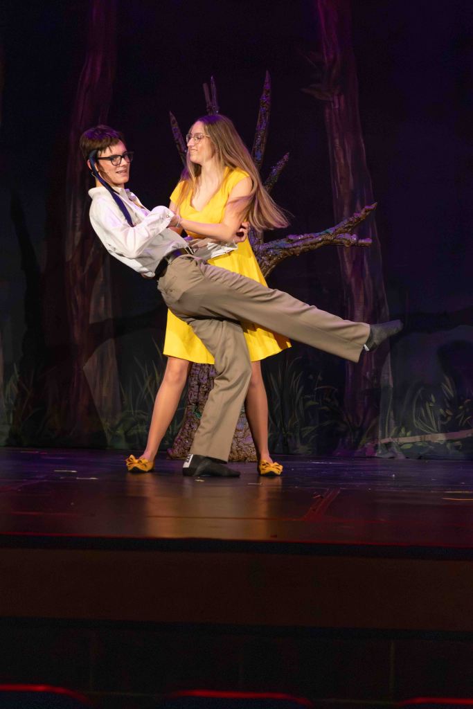 A scene from a performance featuring two actors dancing on stage. One is wearing a white shirt and black suspenders, lifting the other, who is dressed in a yellow dress.