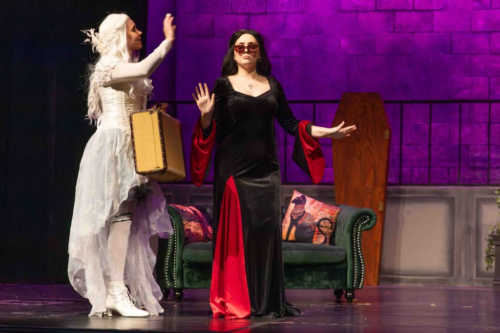 Two actresses performing on stage, one dressed in a flowing white costume and the other in a black and red gown, with dramatic gestures. The stage features a green couch and purple backdrop.