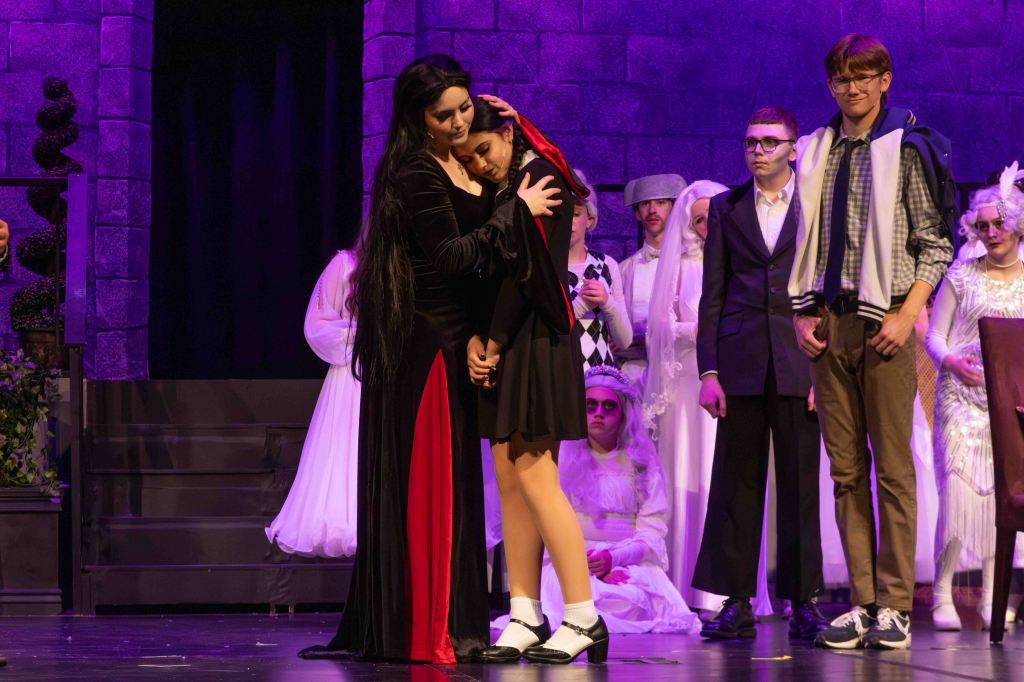 A theatrical scene depicting two female characters in an emotional embrace on stage, with other cast members in the background wearing various costumes, set against a purple-lit backdrop.