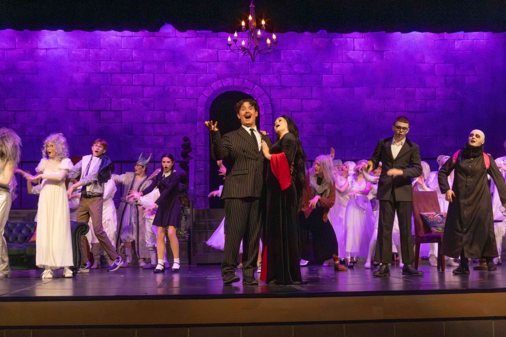 A theatrical performance scene featuring a large cast of actors dressed in various costumes on stage, with purple lighting and a castle-like backdrop. The main characters are singing at the forefront, surrounded by other performers in ghostly attire.