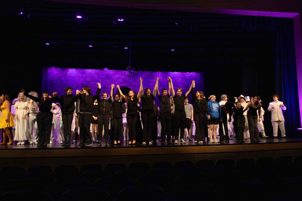 Theater cast members celebrating on stage after a performance, holding hands and raising them in triumph, with a colorful backdrop and audience seats visible.