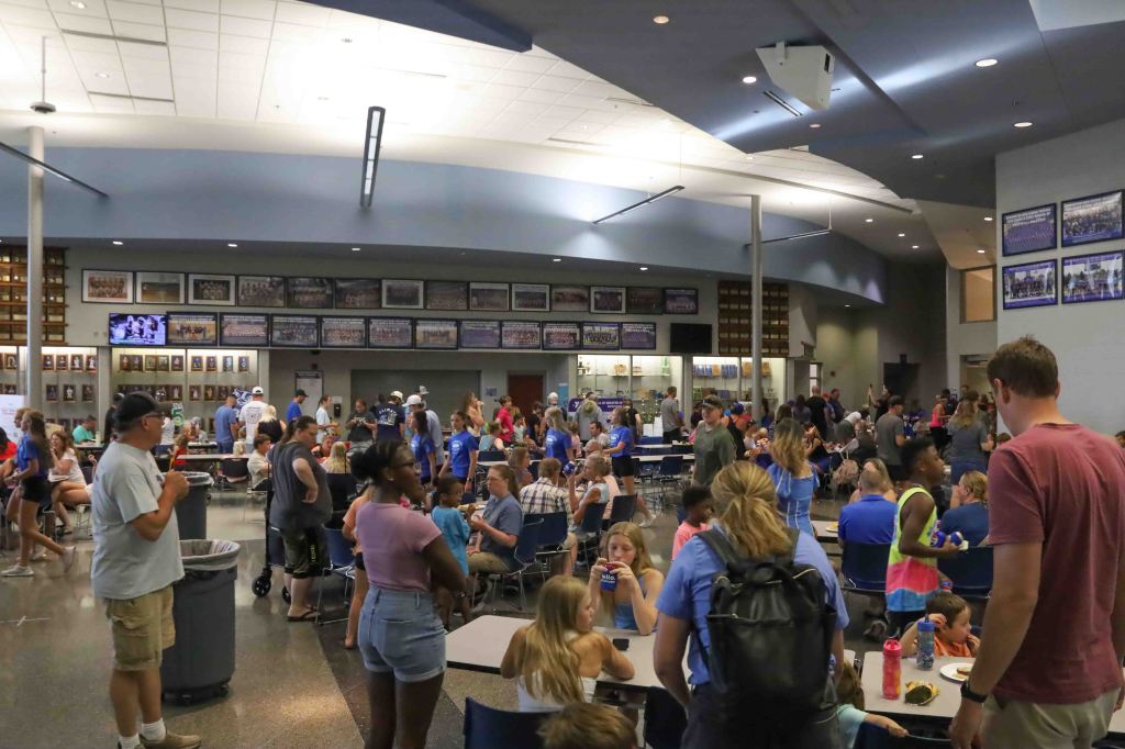 A busy cafeteria filled with people sitting at tables, chatting, and eating. The walls are adorned with framed photos and memorabilia, creating a lively atmosphere.