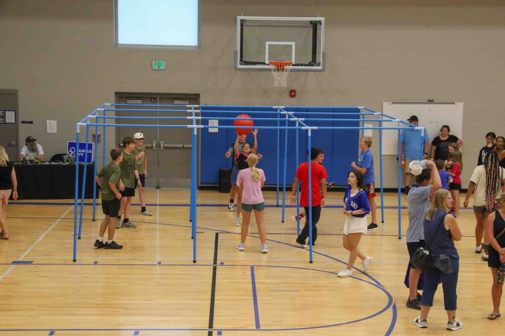 A gymnasium scene with children and adults participating in a group activity. There is a blue framework set up in the center, where several kids are playing with a large red ball. Others are watching and engaging from the sidelines.