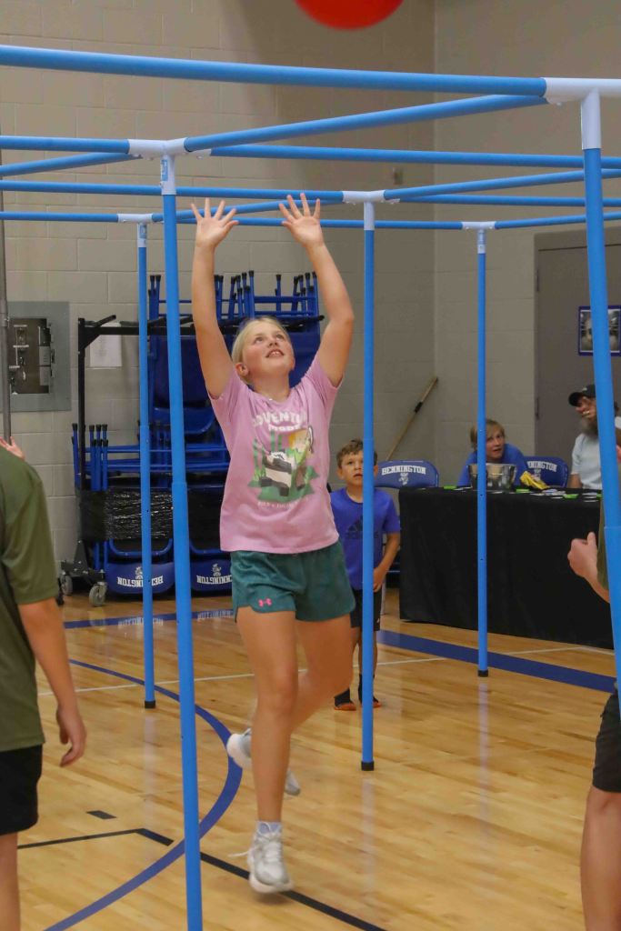 A young girl is reaching up to catch a red ball while playing in a gymnasium, surrounded by other children and blue equipment.