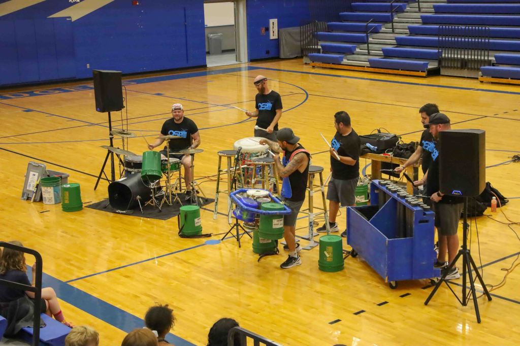 A group of six musicians performs in a gymnasium, using trash cans and other unconventional items as percussion instruments. The floor is wooden, with blue bleachers in the background. Some audience members are seated and watching the performance.