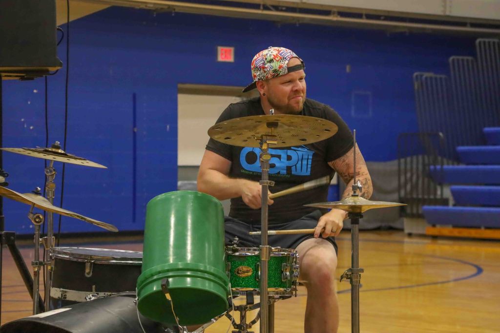 A drummer performing on a makeshift drum set, using a green bucket as a drum and seated in a gymnasium. The drummer wears a colorful cap and a black t-shirt with a graphic design.