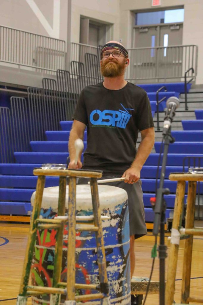 A man playing a drum in a gymnasium, wearing a black t-shirt with 'OSP' printed on it, and a headband, with a focused expression.