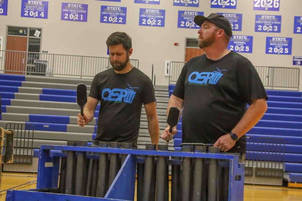 Two men standing in a gymnasium, each holding a microphone, with a blue equipment rack in front of them.