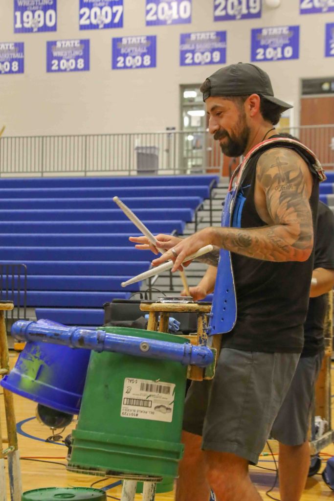A man with tattoos plays percussion on colorful buckets in a gymnasium, wearing a tank top and a cap.