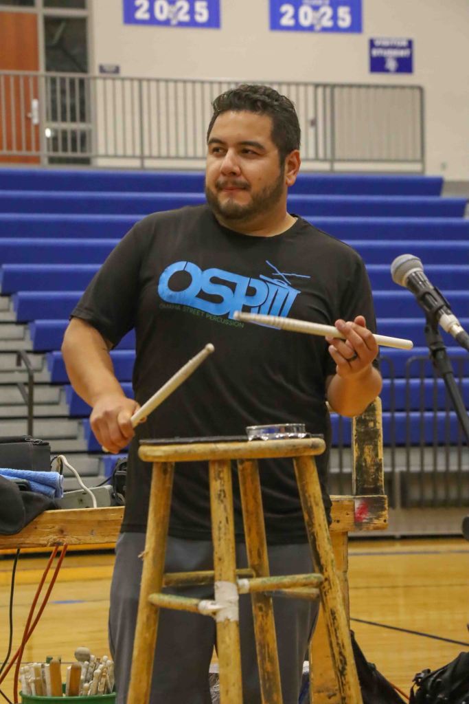 A man stands holding drumsticks, preparing to perform on a wooden stool. He wears a black t-shirt with 'OSP' written on it, and the background features blue bleachers.