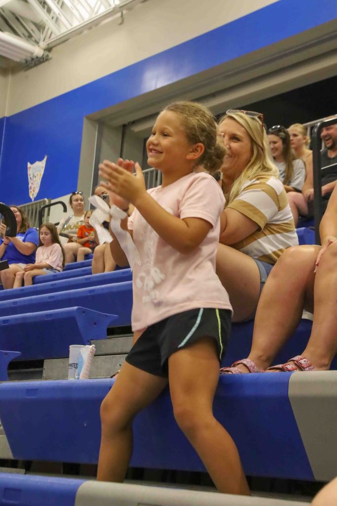 A smiling young girl in a pink shirt and black shorts is standing and clapping in a sports arena, with adults and children seated in the background.