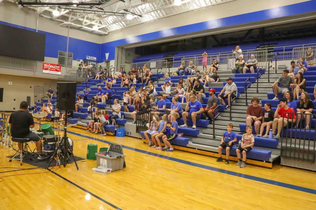 A drummer performs in front of an audience seated in blue bleachers at a gymnasium event, with a variety of people of different ages watching attentively.