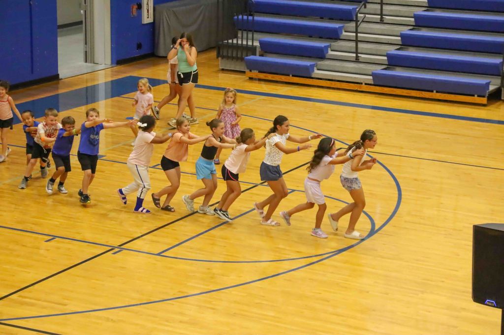 A group of children participating in a line dance activity in a gymnasium, with one adult supervising in the background.