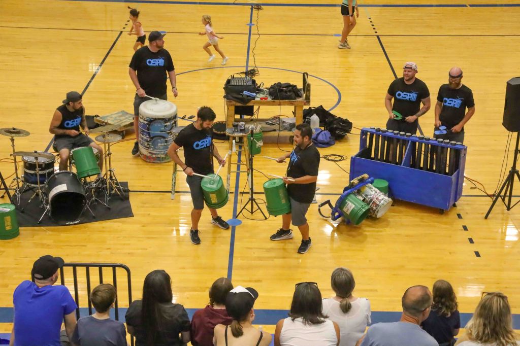A group of musicians performing with various percussion instruments, including drums and buckets, in a gymnasium. Audience members watch from the stands while a child runs in the background.