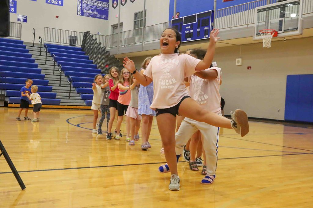 A group of children participating in a fun activity in a gymnasium, with one girl in the foreground joyfully performing a dance move while others stand in a line behind her.